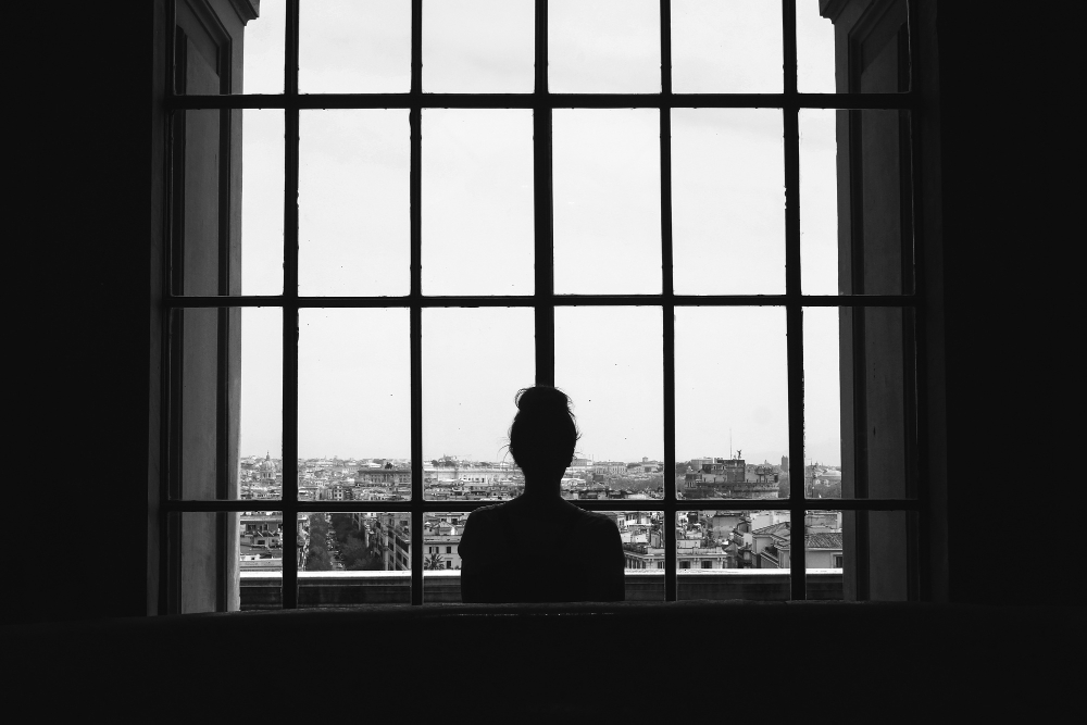 Bidikan hitam putih wanita kesepian berdiri di depan jendela melihat bangunan