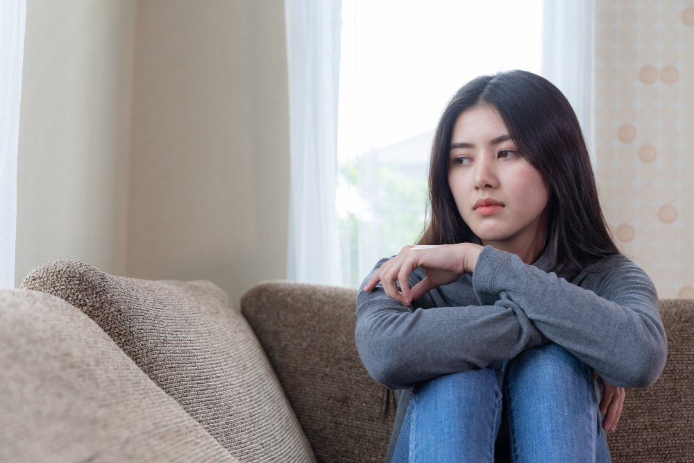 wanita muda cantik asia yang tidak bahagia duduk di sofa sendirian dengan perasaan sedih