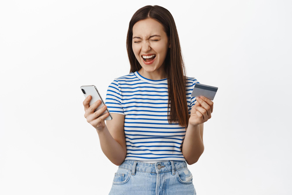 Top Up DANA di Indomaret: excited happy woman scream from happiness winning money cashback holding credit card with mobile phone rejoice standing against white background laughing