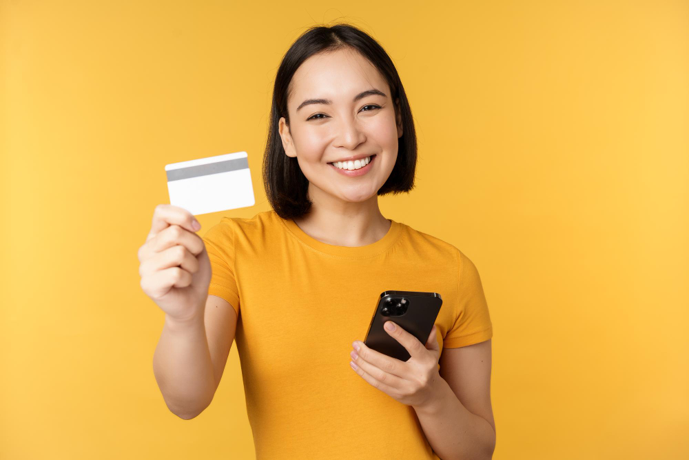 joyful asian girl smiling showing credit card smartphone recommending mobile phone banking standing against yellow background