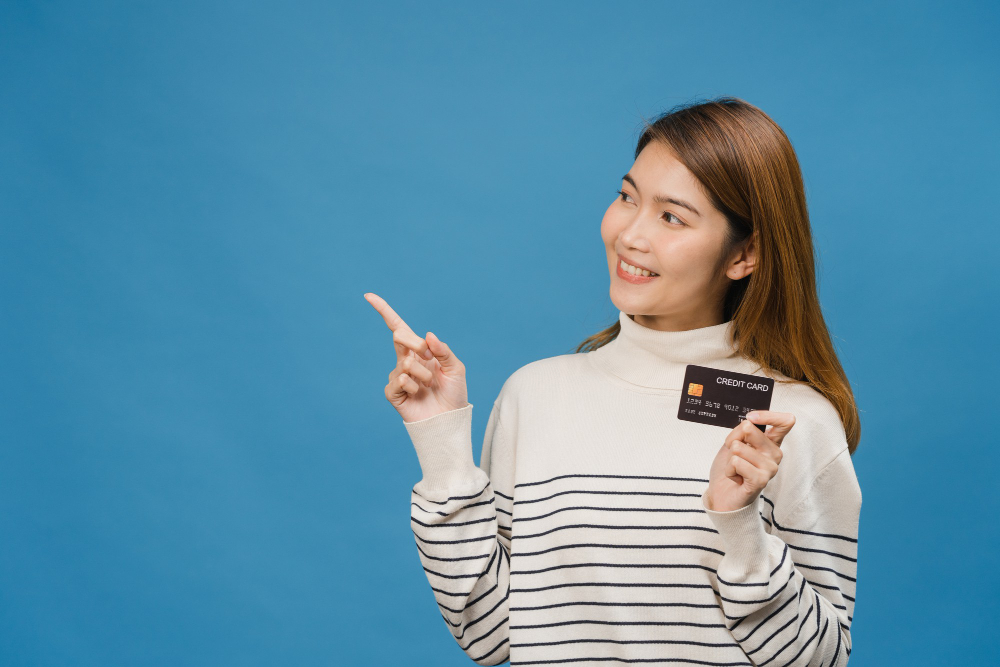 Top Up DANA di Indomaret: young asia lady show credit bank card with positive expression smiles broadly dressed casual clothing feeling happiness stand isolated blue wall